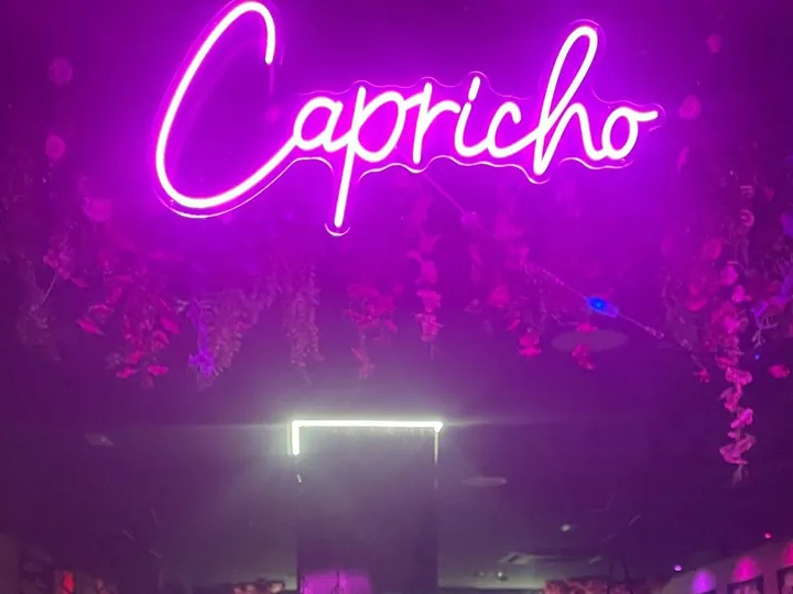 Rótulo de neón rosa con el nombre Capricho en cursiva sobre una pared de flores